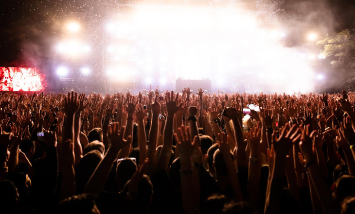 A imagem se trata de um show, onde há uma multidão de braços erguidos de frente para o palco onde se têm um clarão de luzes.