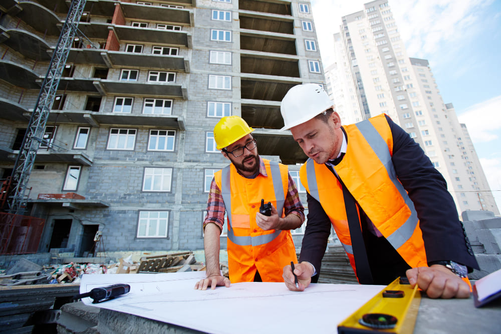 Para poder retratar o Seguro de Riscos de Engenharia, a foto tirada é em uma área de construção civil, onde ao fundo se vê o começo da construção de um prédio, à frente em uma mesa improvisada, há dois homens brancos, que parecem conversar sobre algo, o homem á frente veste um terno por baixo do colete de segurança laranja e um capacete branco, na mão esquerda ele segura uma régua, enquanto na direita um lápis.  O rapaz mais ao lado, está vestido uma camisa xadrez por baixo também do colete de segurança, capacete amarelo e um radio de transmissão na mão. 